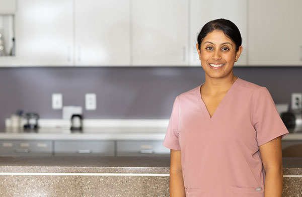 Dr. Neeti Alapati is a woman with brown hair wearing pink scrubs standing in front of a tan desk