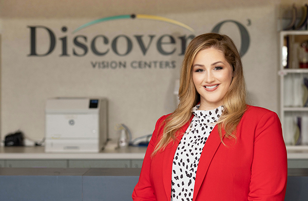 Dr. Jessica Fowler is a caucasian woman standing in front of an offwhite wall with a Discover Vision logo. She is wearing a red suit jacket with a white blouse that has black dots.