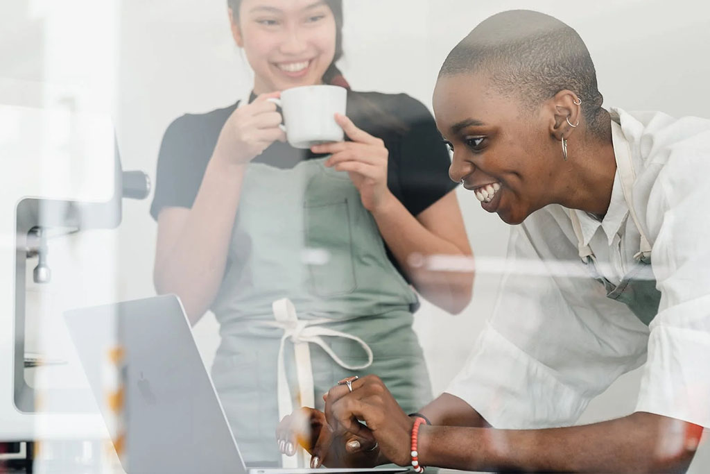 Benefits of laser eye surgery at Discover Vision. Black person with short hair wearing a white shirt looks at their laptop while another person in the background wearing a black shirt and green apron sips coffee.