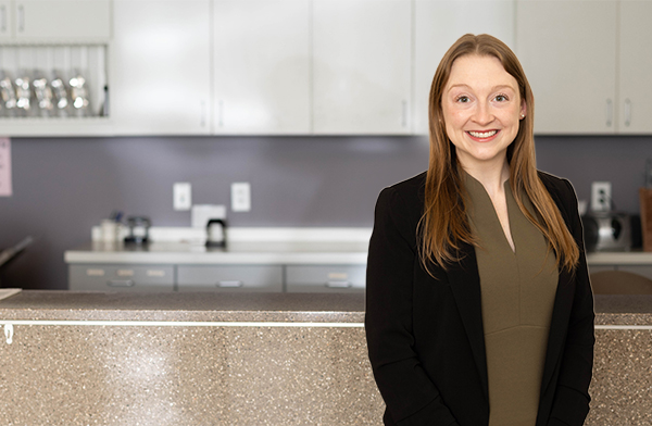 Dr. Elizabeth Bell, OD - Leawood, KS Eye Doctor - caucasian woman with blondish brown hair, wearing a black jacket and sage blouse, stands against a marble counter check-in desk.