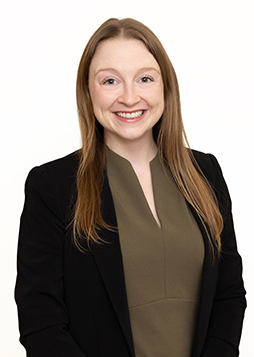 Dr. Elizabeth Bell, OD - a caucasian woman with blondish brown hair stands against a white wall wearing a black jacket and sage-green colored shirt.