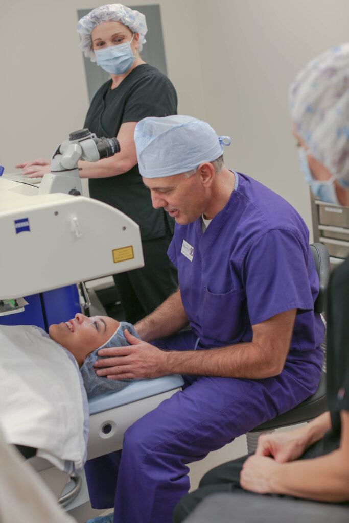 Dr. John Doane, a caucasian male wearing purple scrubs and a surgical cap, talks to a female patient on the operating laser. A surgical technican appears in the background wearing black scrubs and a surgical cap.