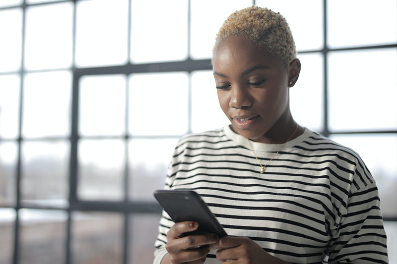 Myopia Symptoms - Black woman with short blonde hair in a striped shirt looks at her cell phone.