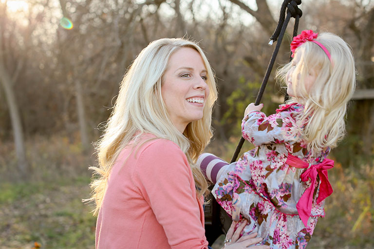 Myopia Treatment - Caucasian woman with blonde hair in a pink sweater pushes her child wearing a floral dress on a swing.