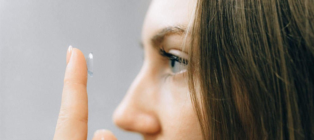 Contact Lens Tips: Caucasian woman with dark hair holds a contact lens on her finger in front of her eye.