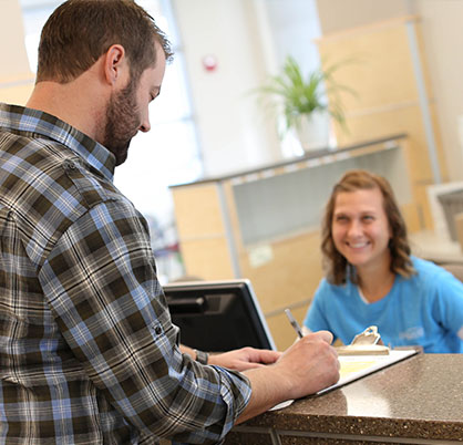 Schedule LASIK appointment. Man with dark hair wearing a plaid shirt checks into the office. Woman with light brown hair, wearing a blue t-shirt smiles from behind the check-in desk.