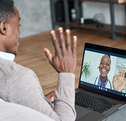 Schedule a LASIK Consult, Virtual. Man waves at laptop screen during virtual appointment and doctor waves back at him on screen.